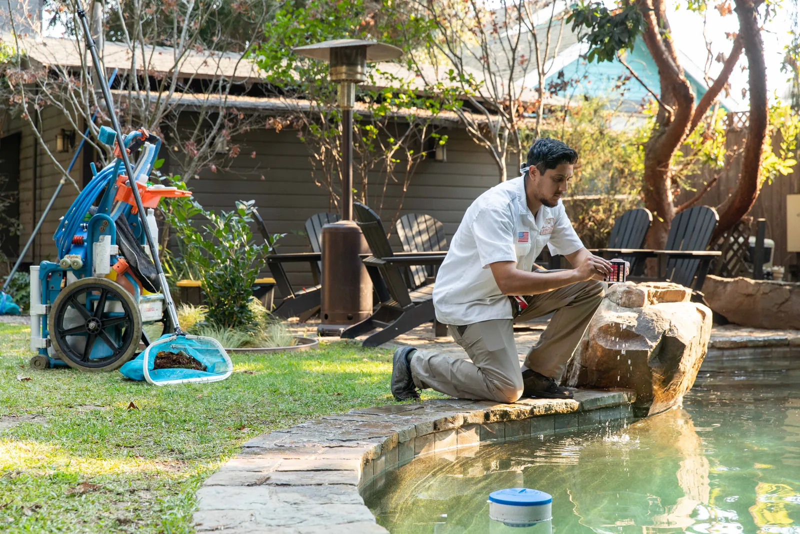 Man crouching at the edge of a pond with a crocodile, trees and outdoor structures in the background.