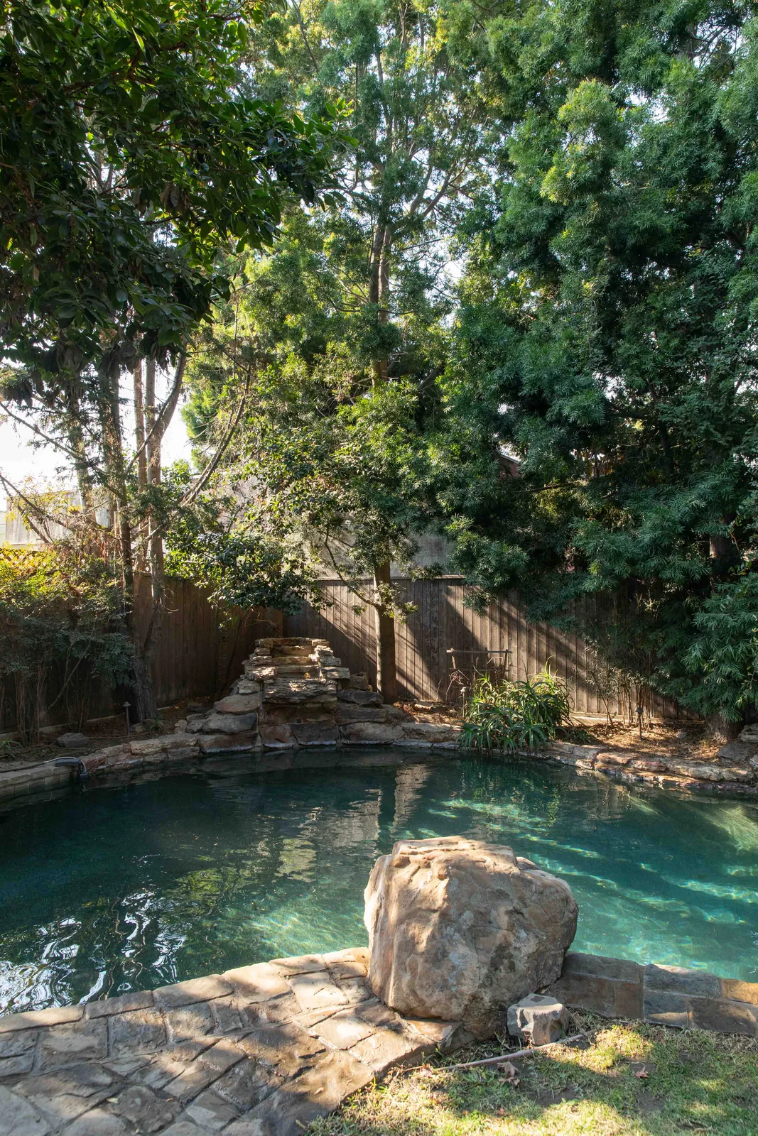 Turquoise pool with a large rock in the center, ringed by dense green trees and dappled sunlight.