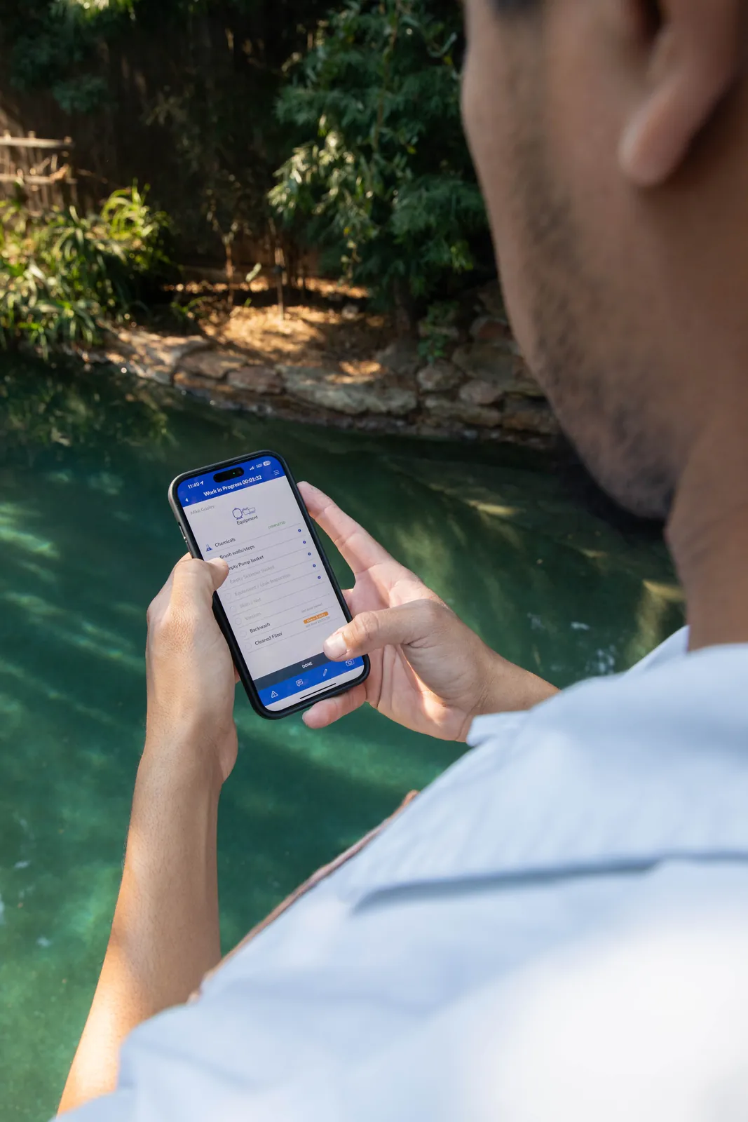 Person from behind holding a smartphone, viewing a bright green pool with trees and foliage in the background.