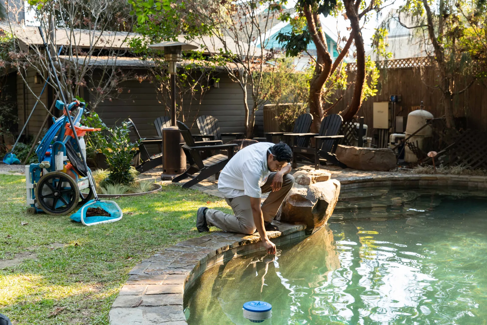 Person bending over the edge of a clear water pool, interacting with the water in a sunny backyard.