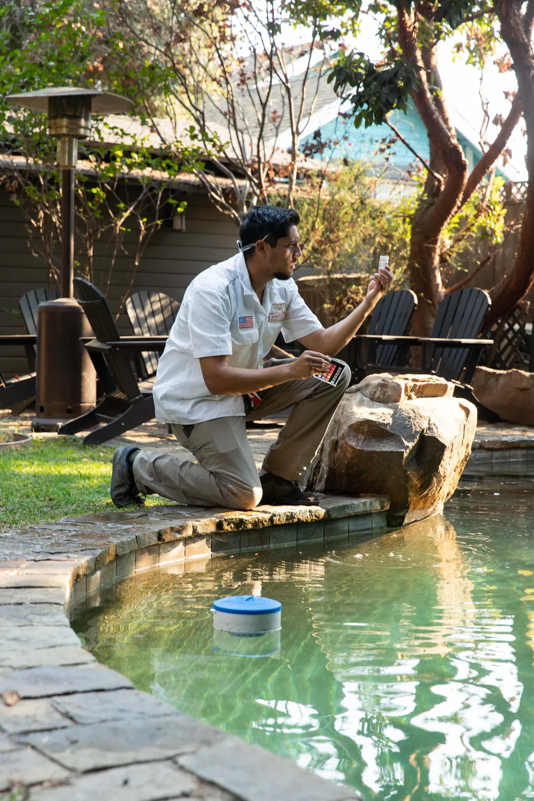 Person crouching beside a rectangular water basin in a garden with large trees and dappled sunlight.