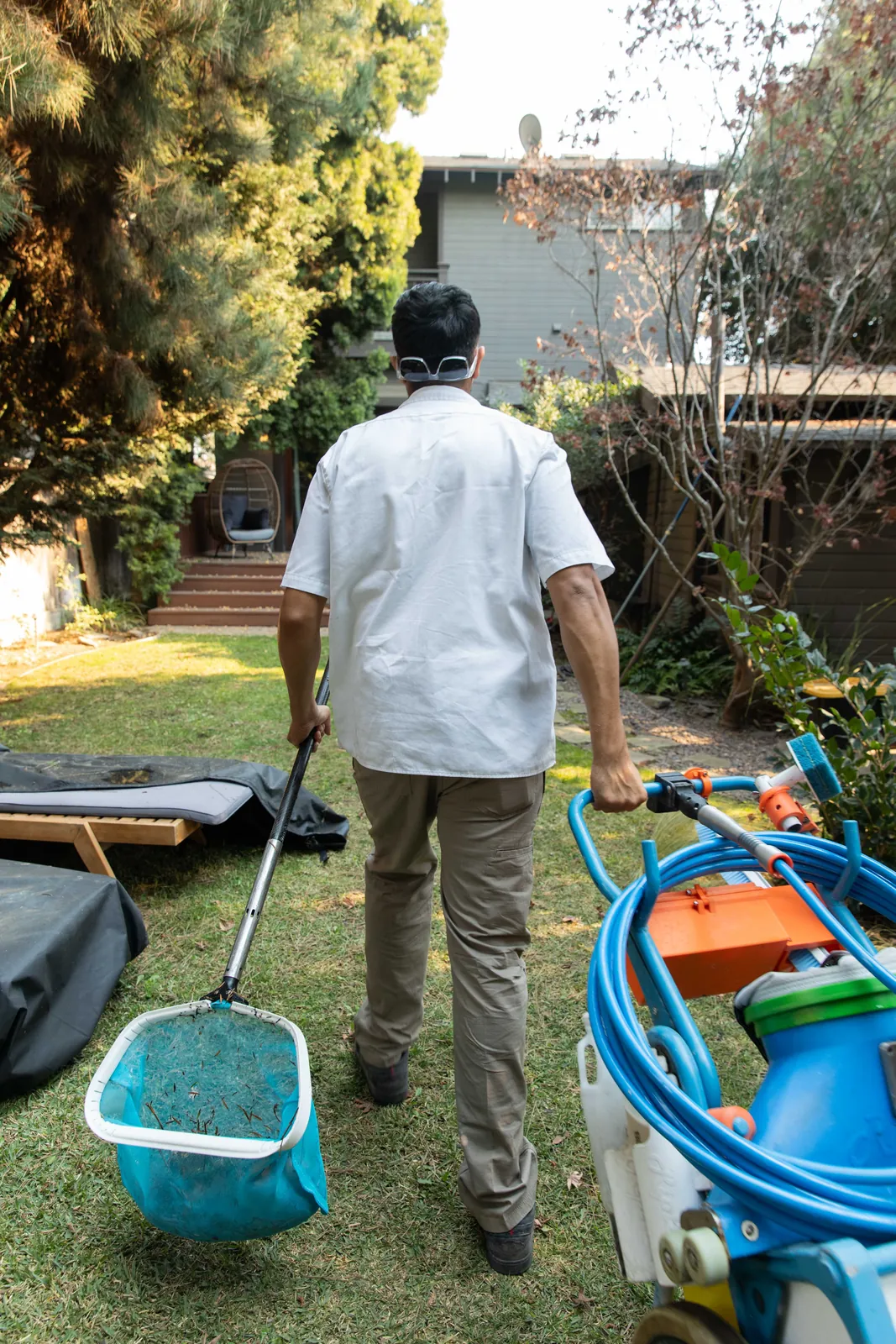Person in white shirt walking past pool maintenance tools and blue equipment in residential yard.
