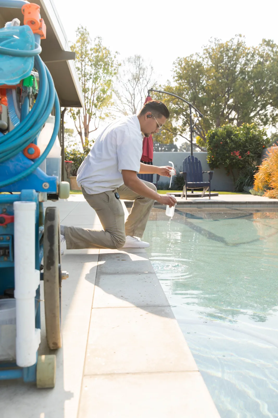 Person in white shirt leaning over pool edge, holding water testing equipment near blue pump system.