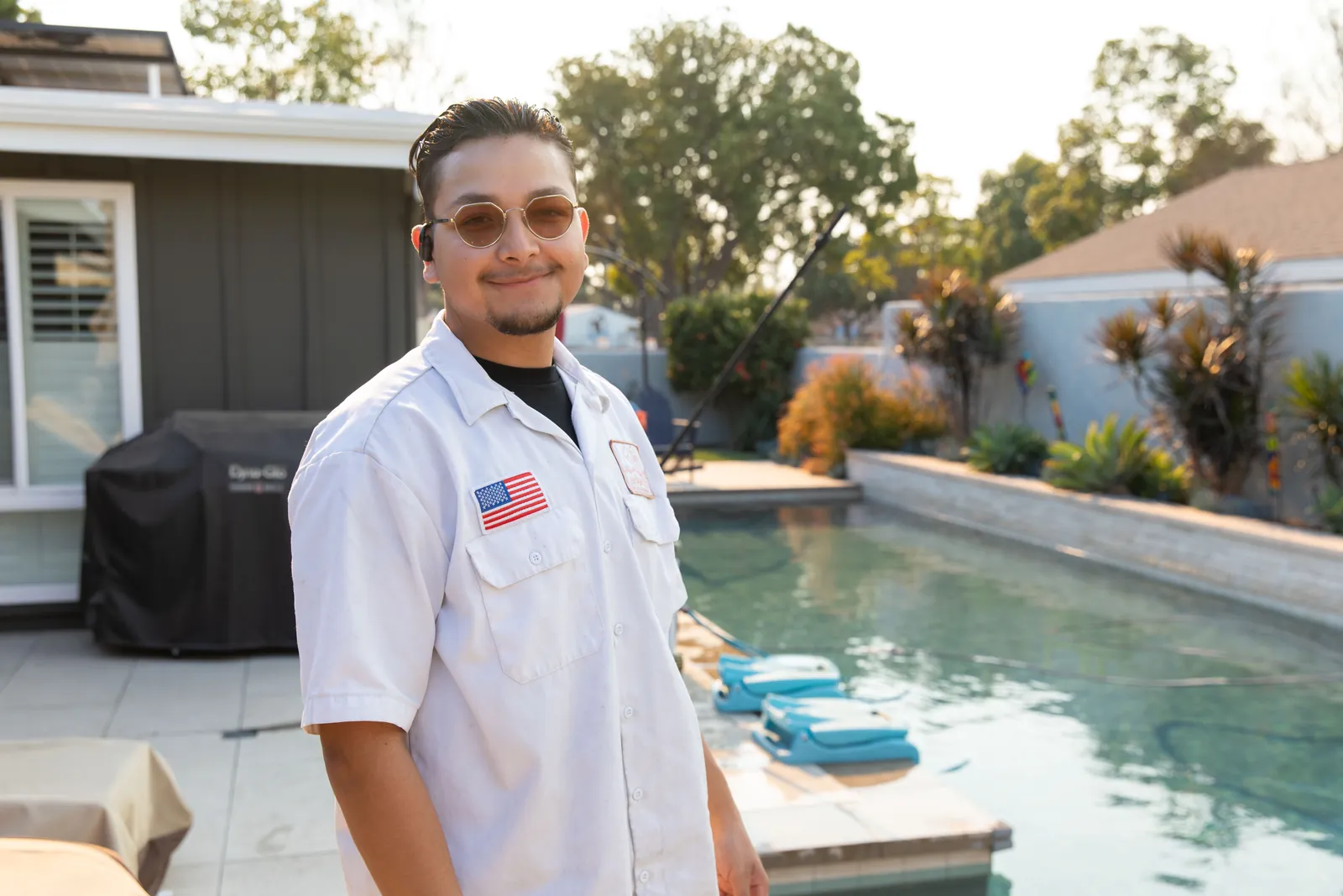 Person in white uniform shirt standing by a residential swimming pool on a sunny day.