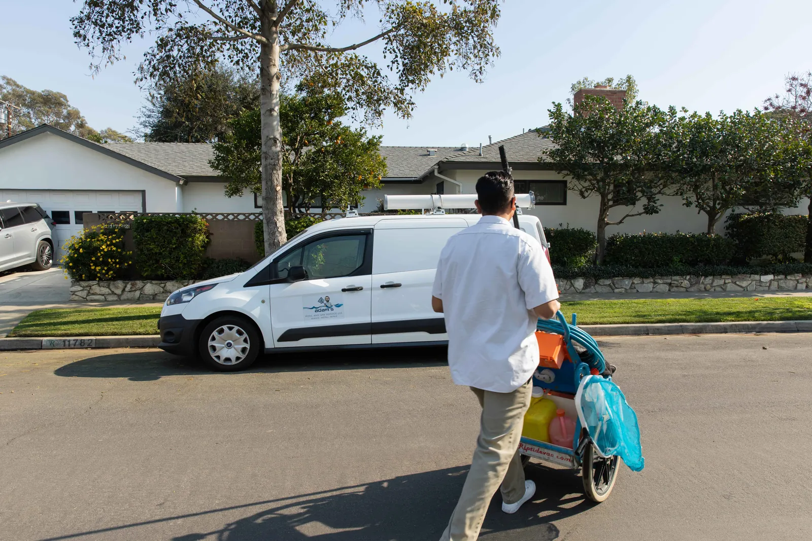 Technician in white shirt holding pool cleaning tools next to white service van on suburban street.