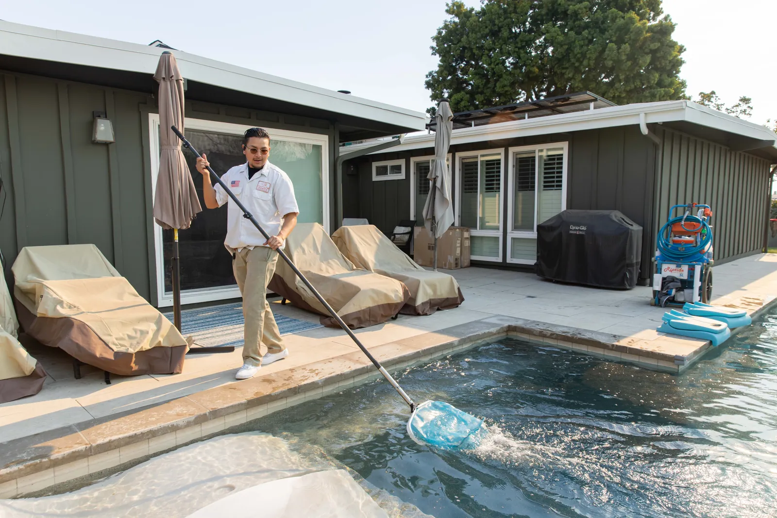Person skimming debris from a pool with lounge chairs and a small pool house visible nearby.